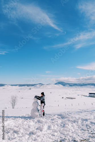Mom and little girl are making a snowman, standing on a snowy clearing in a mountain valley. Back view