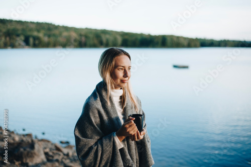 Beautiful young woman relaxing over a cup of tea. A beautiful blonde woman drinks a hot drink from a dark mug in an autumn park close to lake on the sunset. The woman is wrapped in a warm grey scarf.