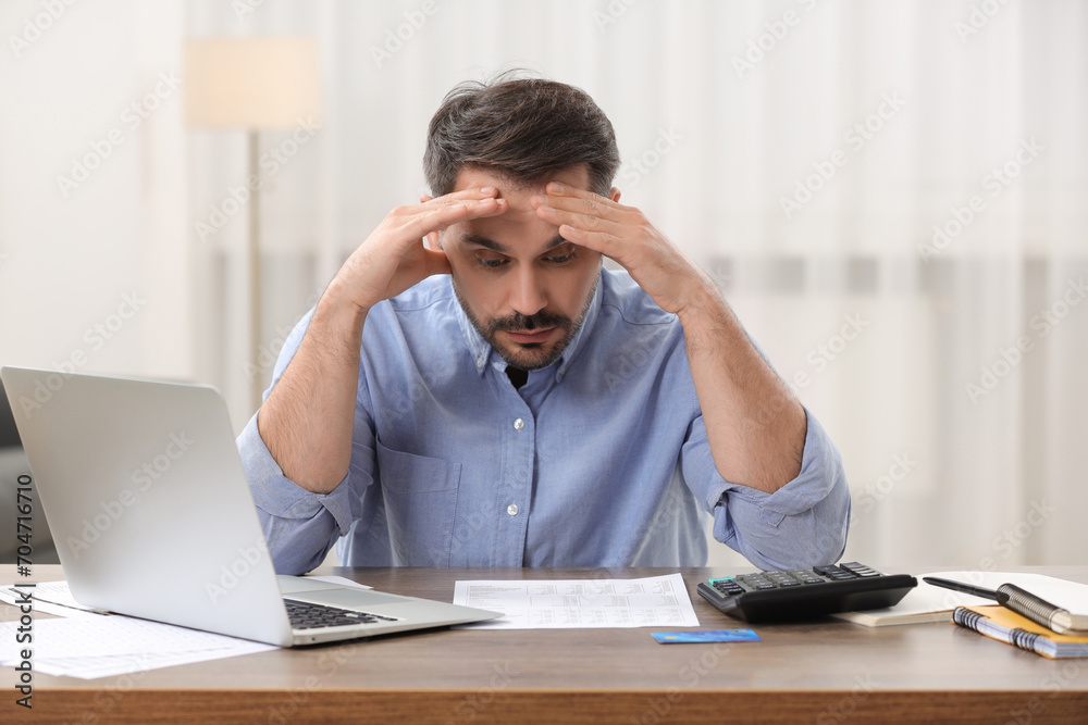 Confused man with debt notification, laptop and credit card planning budget at table in room. Financial problem Confused man with debt notification, laptop and credit card planning budget at table in room. Financial problem