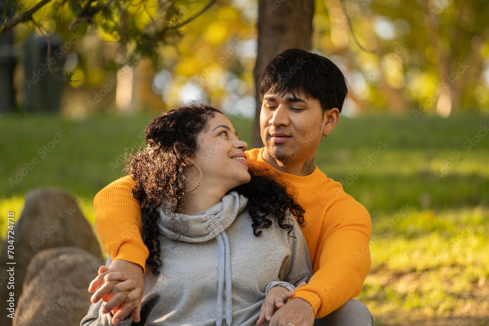 Fototapeta premium Latin couple hugging and flirting in an urban park sitting in the grass and looking each other