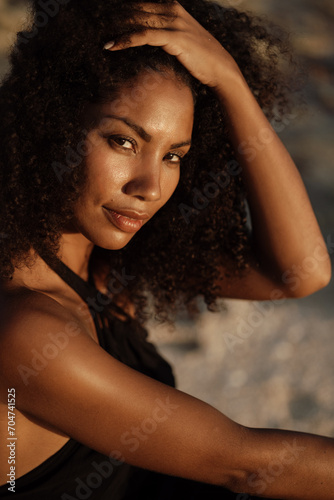 Beautiful young african american girl sitting on sand at the beach and look at the camera. African girl with curly hair look at the camera, nature background