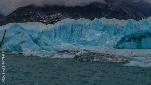 A melting glacier. A wall of blue ice rises above a turquoise glacial lake. Thawed ice floes, broken-off icebergs float in the water. Mountains in clouds and fog. Argentina. El Calafate. Patagonia.