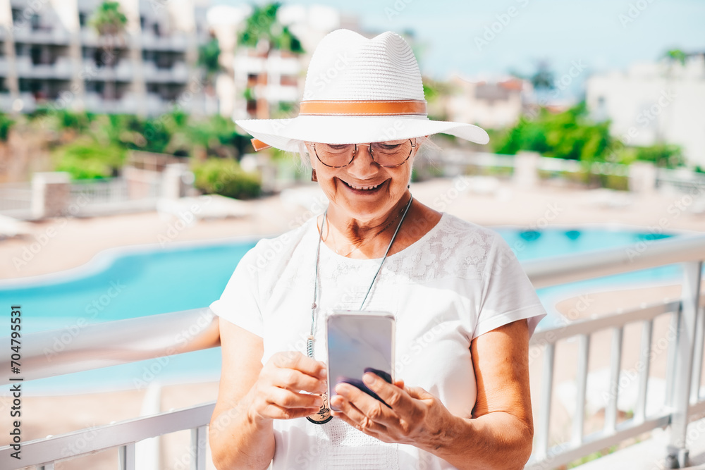 Smiling attractive senior woman with white hat using phone typing or reading a message standing outdoor under the sun Smiling attractive senior woman with white hat using phone typing or reading a message standing outdoor under the sun