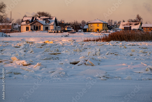 Winter landscape with frozen river with ice drifts and houses at sunset. Landscape with golden sunset light in cold winter evening in Latvia. Jelgava town.