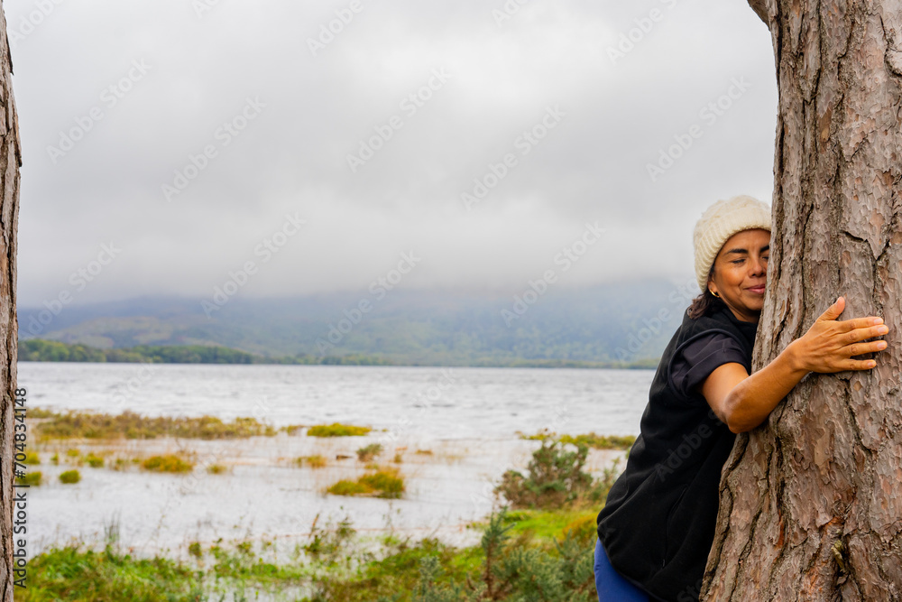 Colombian woman enjoying a moment of connection in nature on her trekking route through Ireland trying to hug a large tree