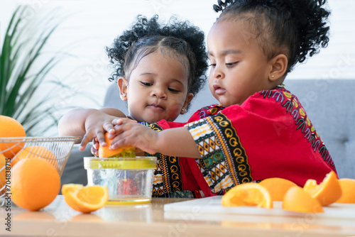 Two African cute kid girls squeezing fresh oranges at home. Adorable children siblings help making freshly squeezed orange juice on manual juicer together. Healthy lifestyle and learning concept photo