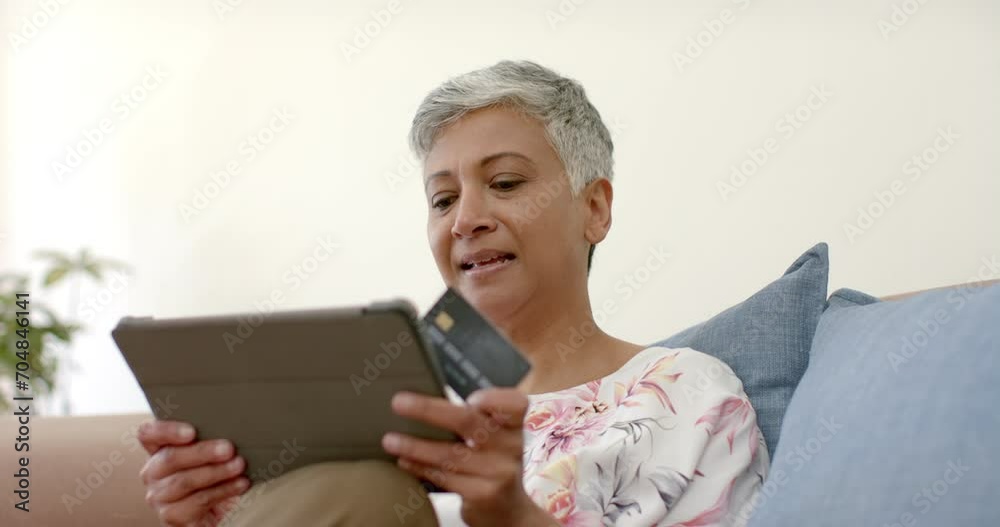 Happy senior biracial woman on couch holding credit card and using tablet at home, slow motion Happy senior biracial woman on couch holding credit card and using tablet at home, slow motion