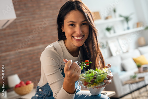 Pretty smiling woman eating healthy salad while sitting on the kitchen table at home.
