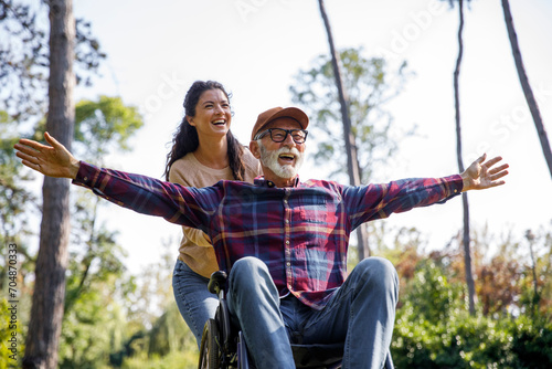 Social worker pushing cheerful retired senior man with arms outstretched in wheelchair at park photo
