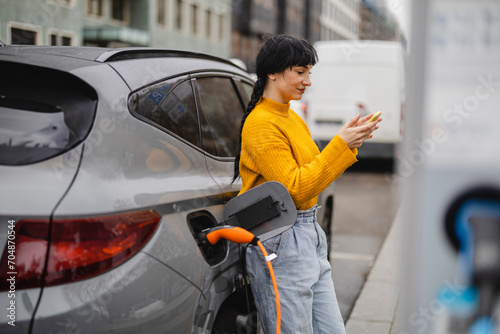 Woman using smart phone and leaning on car at electric vehicle charging station photo