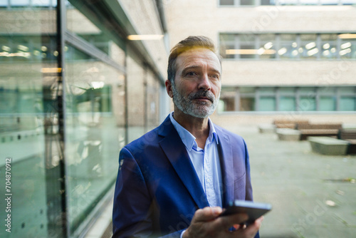 Thoughtful businessman holding smart phone seen through glass photo