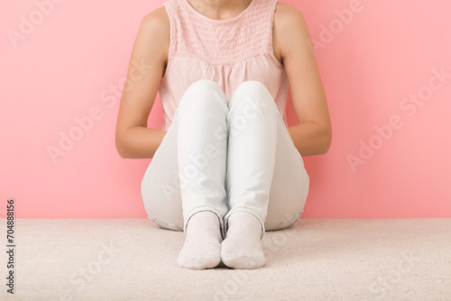 Alone girl in shirt, white jeans and socks sitting on light beige carpet at pink wall background. Legs closeup. Pastel color. Front view. photo
