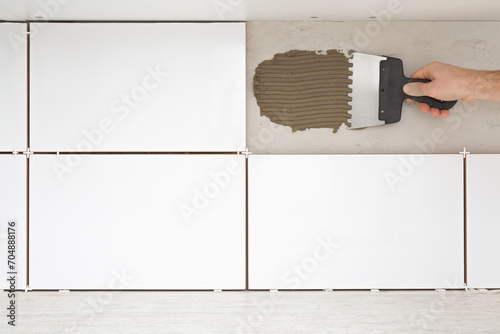 Young man hand using spatula and putting fresh glue on wall concrete between kitchen cabinets and table top. Gluing white ceramic tiles. Closeup. Front view. Renovation process. Repair work of home. photo