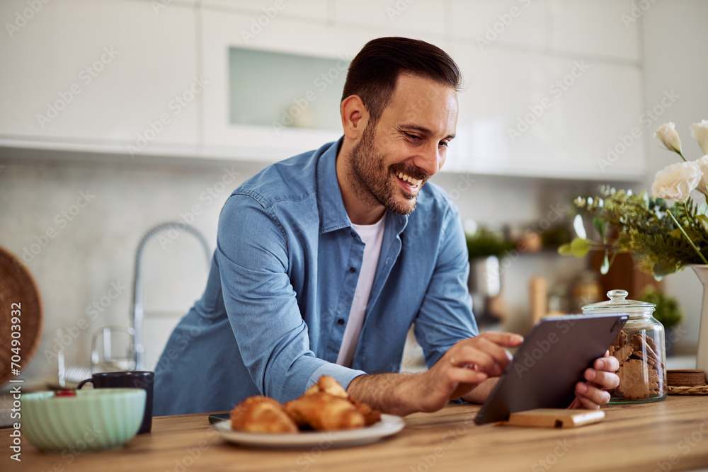 A cheerful man answers a video call on a tablet while leaning on a kitchen counter. A cheerful man answers a video call on a tablet while leaning on a kitchen counter.