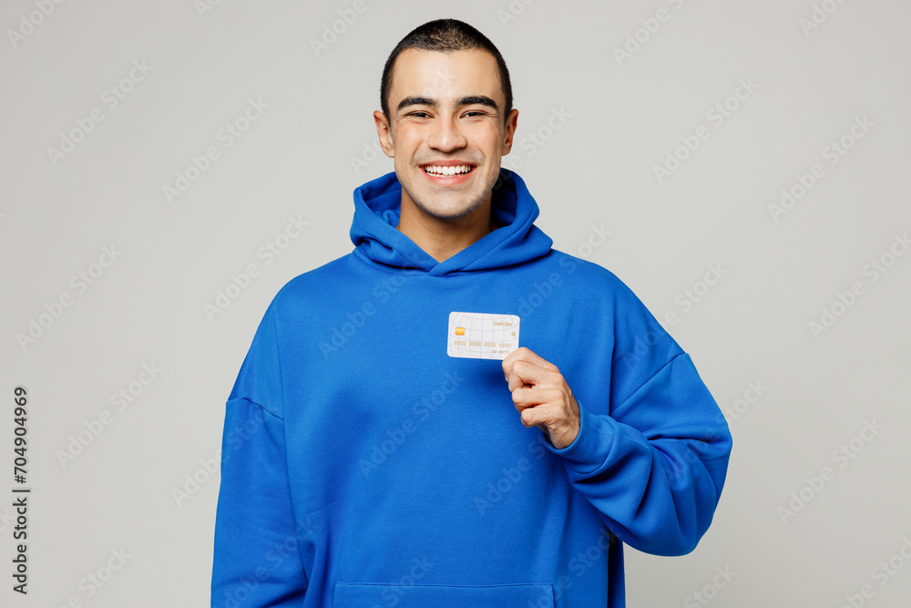 Young smiling happy middle eastern man wear blue hoody casual clothes hold in hand mock up of credit bank card look camera isolated on plain solid white background studio portrait. Lifestyle concept. Young smiling happy middle eastern man wear blue hoody casual clothes hold in hand mock up of credit bank card look camera isolated on plain solid white background studio portrait. Lifestyle concept.