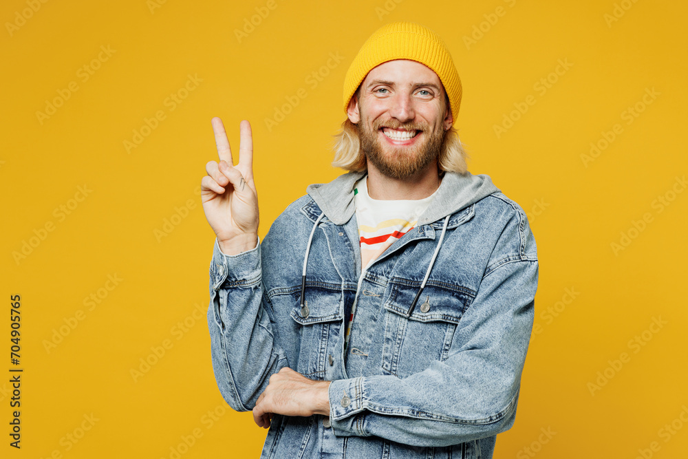 Young smiling happy cheerful fun blond man he wears denim shirt hoody beanie hat casual clothes showing victory sign look camera isolated on plain yellow background studio portrait. Lifestyle concept.
