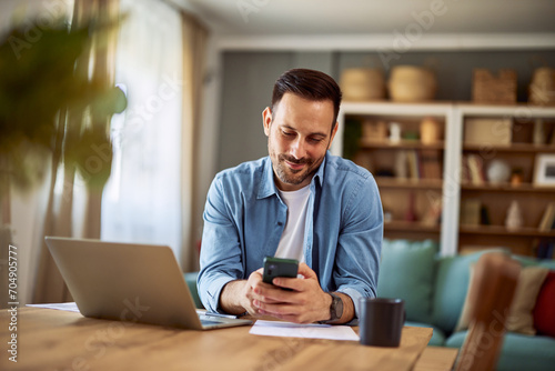 Wall Mural A smiling man checking his messages on his smartphone while sitting at a table with a laptop in front of him