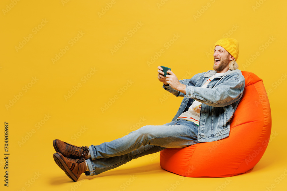 Full body young man wear denim shirt hoody beanie hat casual clothes sit in bag chair use play racing app on mobile cell phone gadget smartphone for pc video games isolated on plain yellow background