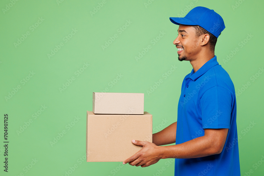 Sideways happy delivery guy employee man wears blue cap t-shirt uniform workwear work as dealer courier hold stack of blank cardboard box package isolated on plain green background. Service concept. Sideways happy delivery guy employee man wears blue cap t-shirt uniform workwear work as dealer courier hold stack of blank cardboard box package isolated on plain green background. Service concept.