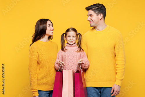 Young happy smiling parents mom dad with child kid girl 7-8 years old wear pink sweater casual clothes hold shopping package bags isolated on plain yellow background Black Friday sale buy day concept