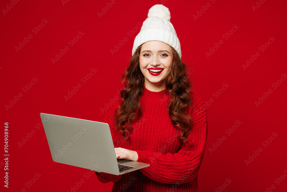 Young happy smart smiling fun IT woman she wears knitted sweater white hat casual clothes hold use work on laptop pc computer isolated on plain red color background studio portrait. Lifestyle concept. Young happy smart smiling fun IT woman she wears knitted sweater white hat casual clothes hold use work on laptop pc computer isolated on plain red color background studio portrait. Lifestyle concept.