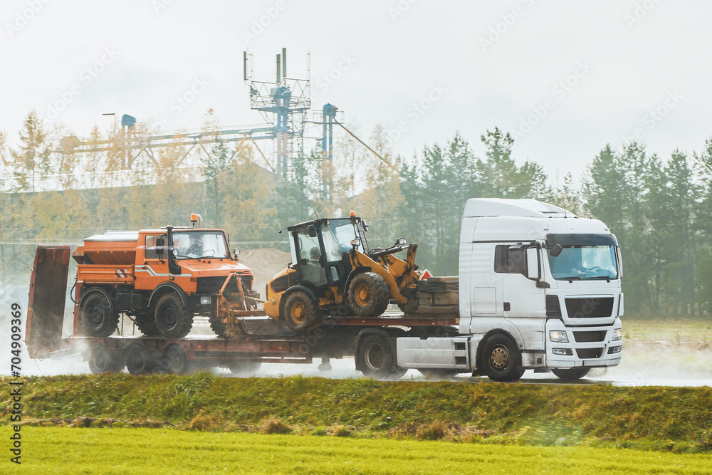 Fototapeta premium The loader and bulldozer are transported by truck. Heavy construction equipment and trucks on a trailer. Amidst a dusty atmosphere on the highway. White truck transports road working machinery.