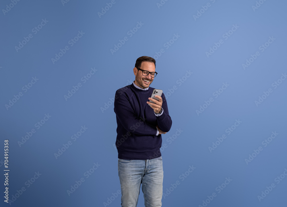 Smiling handsome businessman text messaging over smart phone while standing on blue background Smiling handsome businessman text messaging over smart phone while standing on blue background