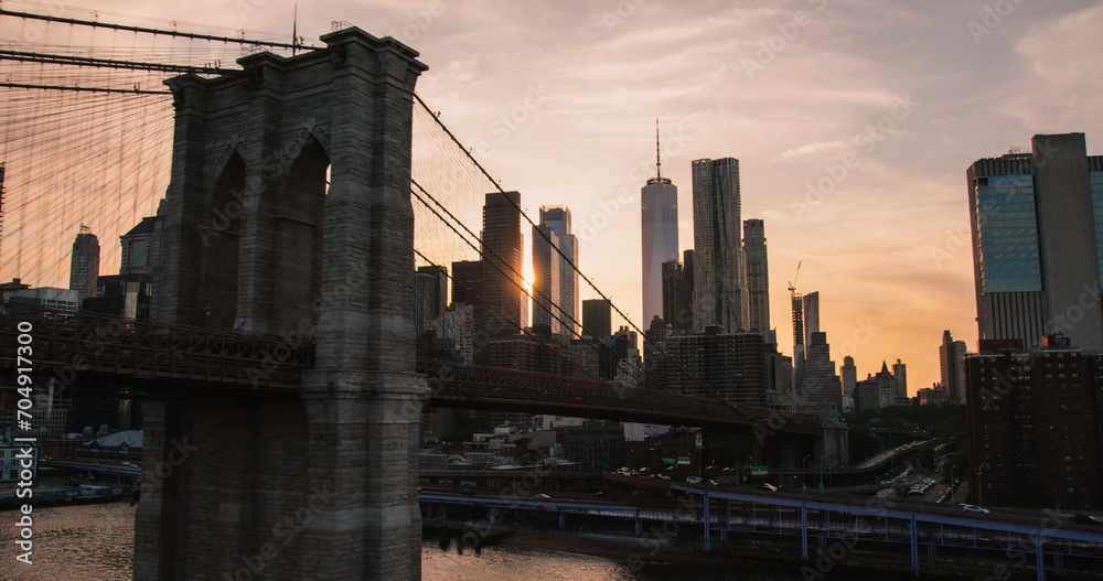Fototapeta premium Aerial Cinematic Drone Scene Near the Brooklyn Bridge with Lower Manhattan Skyscrapers Cityscape. Beautiful Evening Sun Shining with Warm Sunset Light. Shot of a Suspension Bridge Architecture
