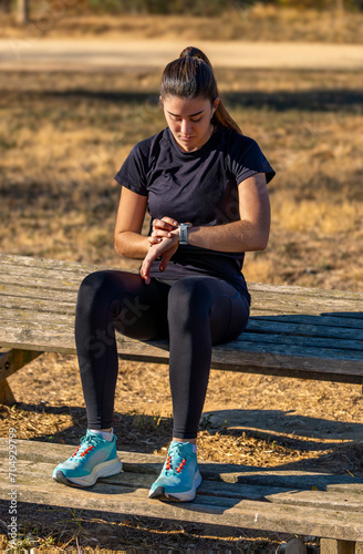 Young runner girl in sportswear sitting on a wooden bench in a park, enjoying listening to music with white wireless headphones looking at her sports results on her smart watch.