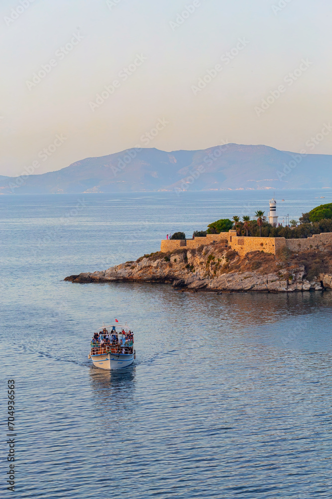 Pigeon Island Castle, (Guvercinada kalesi) and tourist boat at the sunset and gentle pastel sky and mountain silhouette at background. Vertical shot. Kushadasi, Turkey