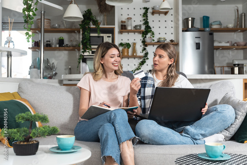 Two young women sitting on the sofa using laptop computer and credit card for online shopping and ordering groceries and food for home so they dont have to go out to buy food photo