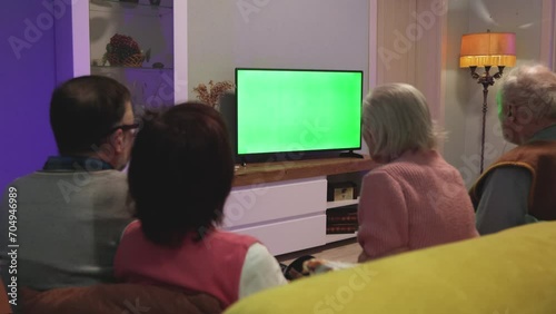 Family watching TV. Green screen. Two couples, an elderly and a middle-aged couple, are sitting on the couch at home. In front of them is a green screen TV. Horizontal panning. 