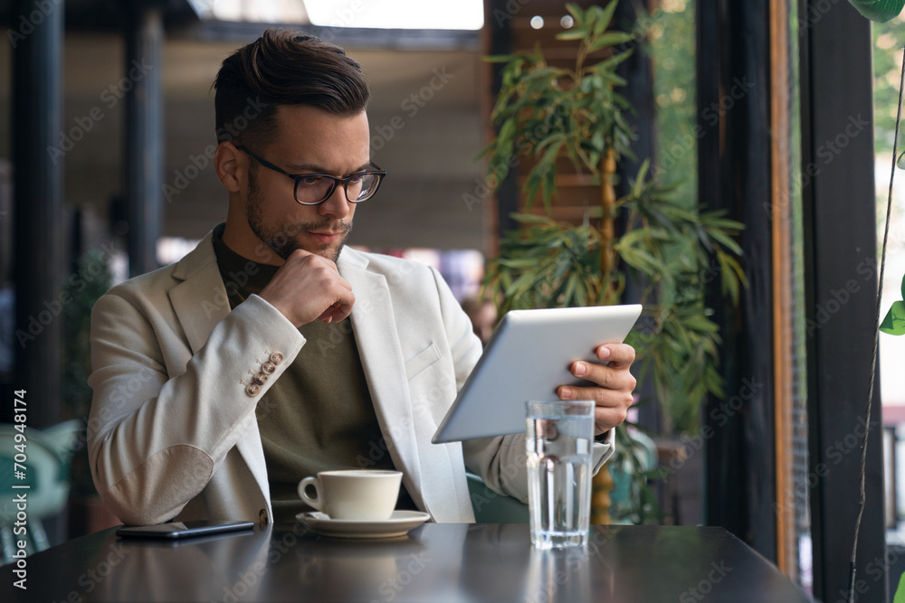 Serious businessman wearing suit and eyewear using digital tablet sitting at table in a Cafe, communicating online, checking emails, distantly working.