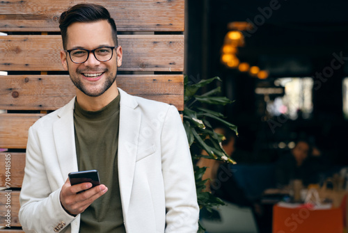 Portrait of young man professional CEO looking at camera and using smartphone while standing in front of restaurant.