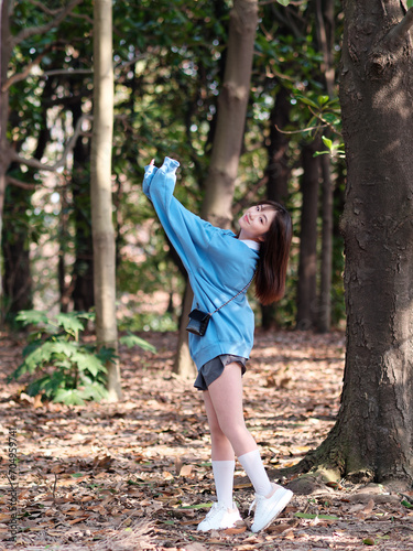 фотографія Beautiful young Chinese woman posing in sunny summer forest, wearing blue loose oversized top and miniskirt with small shoulder bag