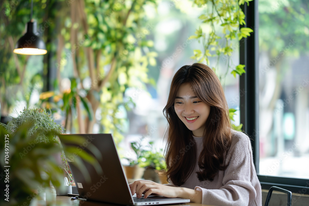 Happy Young Korean Woman Working on Laptop in a Green Cafe Environment Happy Young Korean Woman Working on Laptop in a Green Cafe Environment