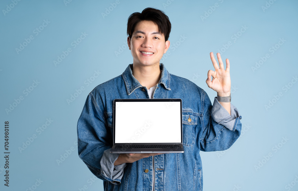 Portrait of an Asian guy wearing a jacket and using a laptop posing on a blue background Portrait of an Asian guy wearing a jacket and using a laptop posing on a blue background