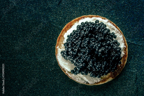 A blini with caviar and cream cheese, overhead flat lay shot on a black slate background with a place for text