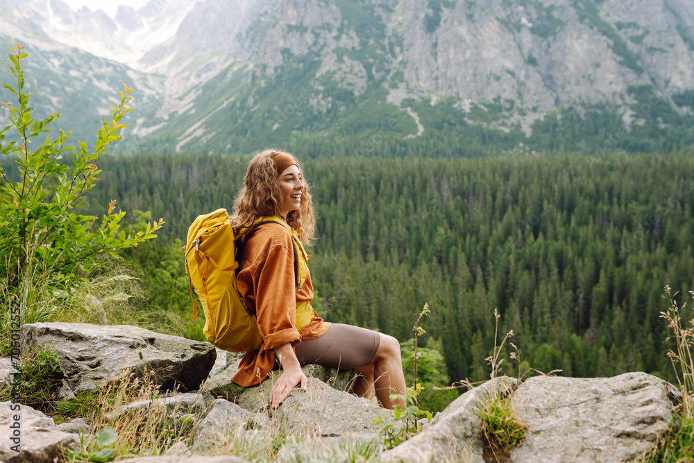 Naklejka premium Happy woman with a yellow hiking backpack enjoying the mountain landscape. A young traveler travels along mountain paths. Adventure, travel concept.