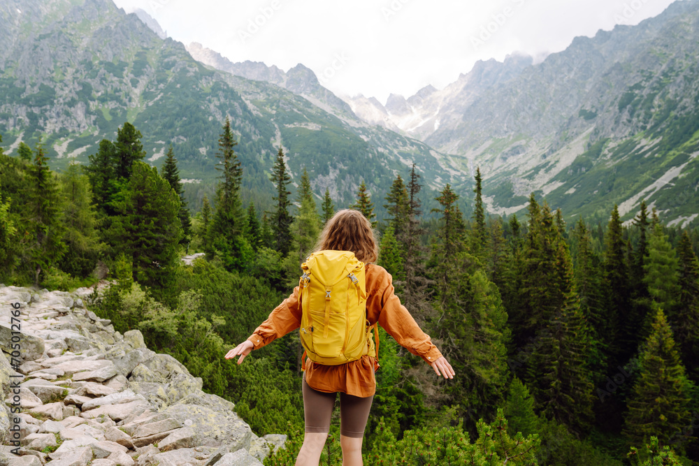 Happy woman with a yellow hiking backpack enjoying the mountain landscape. A young traveler travels along mountain paths. Adventure, travel concept.