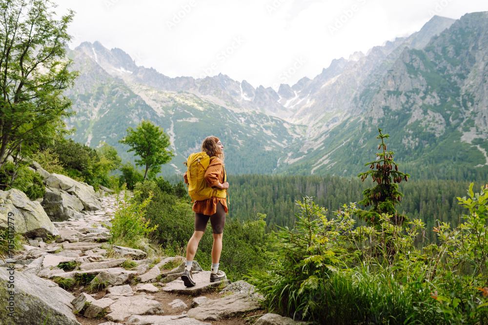 Happy woman with a yellow hiking backpack enjoying the mountain landscape. A young traveler travels along mountain paths. Adventure, travel concept.
