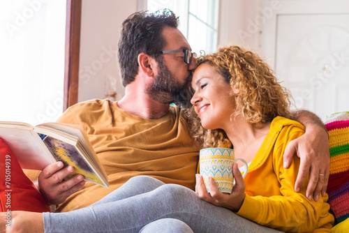 Happy beautiful caucasian couple at home enjoy morning leisure activity in relax together - love and relationship life with young people sitting on the colorful sofa with book and coffee