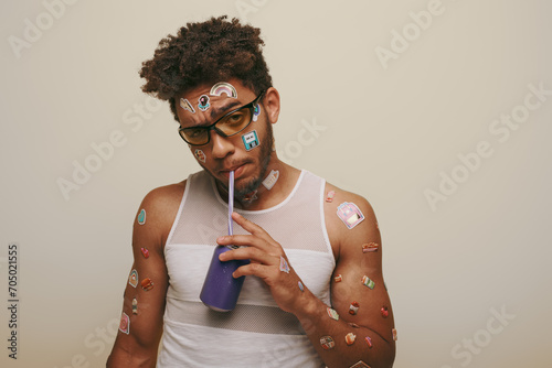 фотографія young african american guy with stickers on face and body drinking soda on grey
