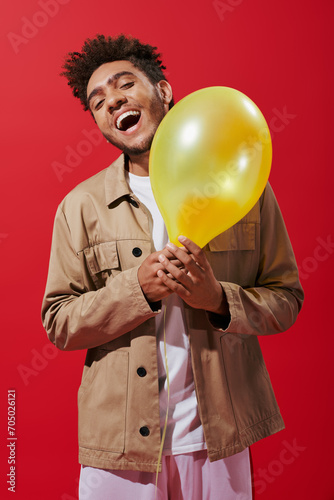 excited african american man in beige jacket holding balloon and smiling on red background © LIGHTFIELD STUDIOS