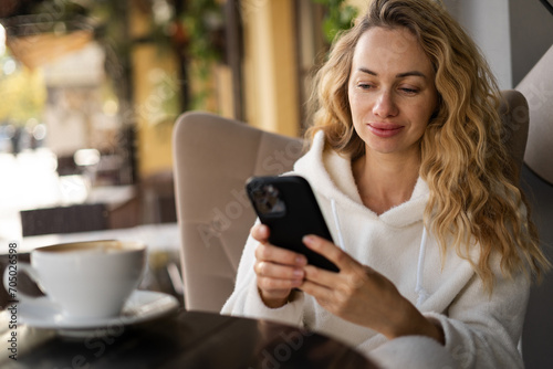 Beautiful blonde woman holding cell telephone typing text message or promotional content, lady watching video on mobile phone during coffee break in cafe or coffee shop photo