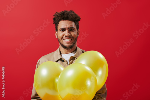 funny african american man in beige jacket holding balloons and grinning on red background © LIGHTFIELD STUDIOS