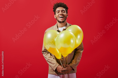 cheerful african american man in beige jacket holding balloons and laughing on red background © LIGHTFIELD STUDIOS