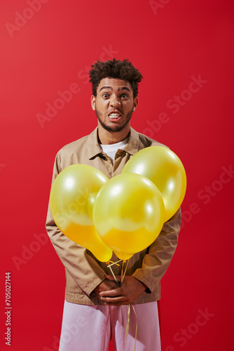 funny african american man in beige jacket holding balloons and grimacing on red background © LIGHTFIELD STUDIOS