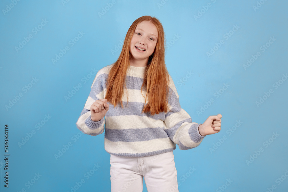 Fototapeta premium Smiling redhead teenage girl dancing on blue background in studio.