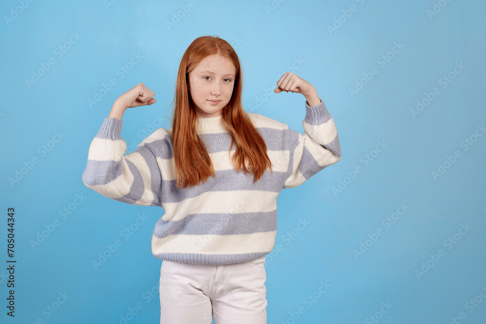 Smiling redhead teenage girl raises arms and shows biceps on blue background in studio. Smiling redhead teenage girl raises arms and shows biceps on blue background in studio.
